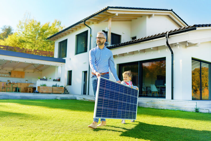 Father With His Little Daughter Carring Solar Panel At Their Backyard. Alternative Energy, Saving Resources And Sustainable Lifestyle Concept.